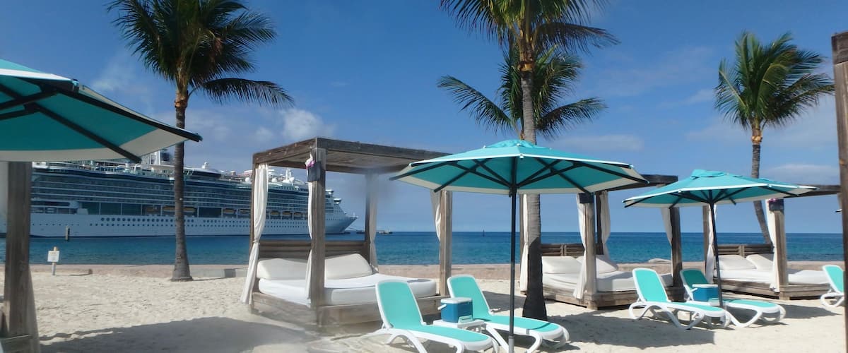 A beach with lounge chairs and umbrellas, palm trees, and a cruise ship in the background.