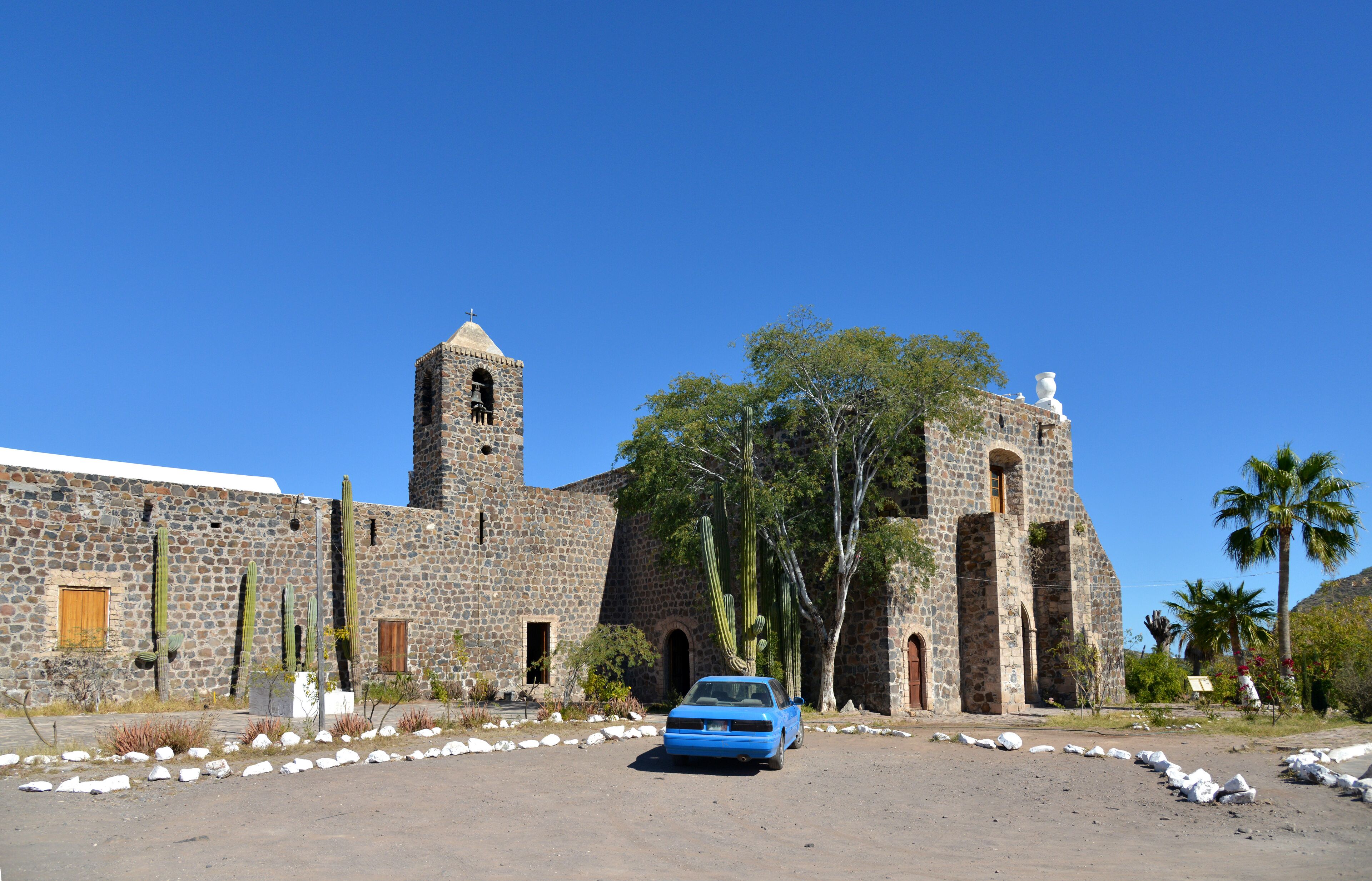 A historic stone building with a bell tower and a blue car parked in front.