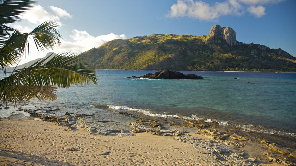 A tropical beach with clear turquoise water, a rocky shoreline, and a prominent mountain in the background.