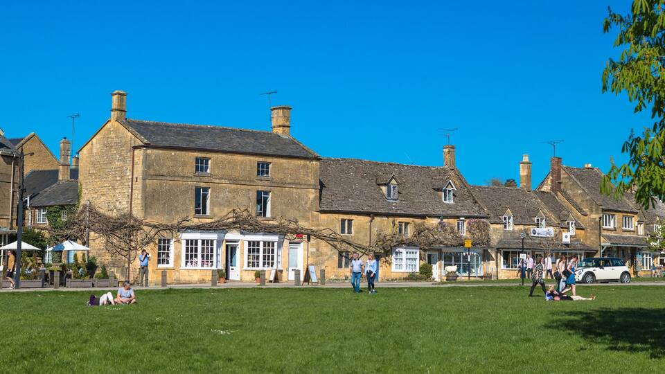 A historic town square with traditional stone buildings, a grassy lawn, and people walking.