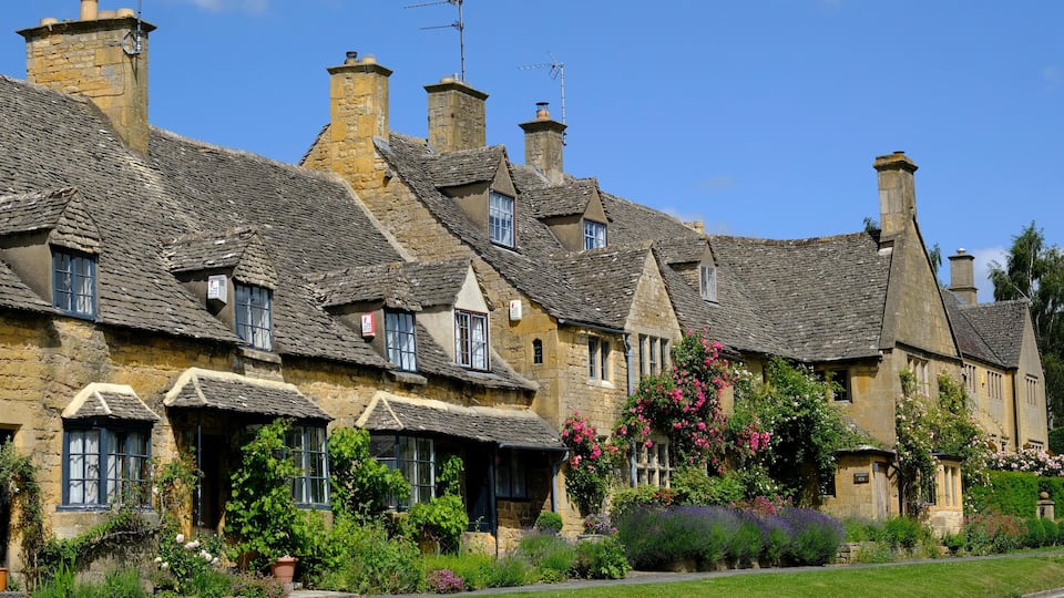 A row of stone houses with steeply pitched roofs and chimneys, surrounded by lush gardens and flowering plants.