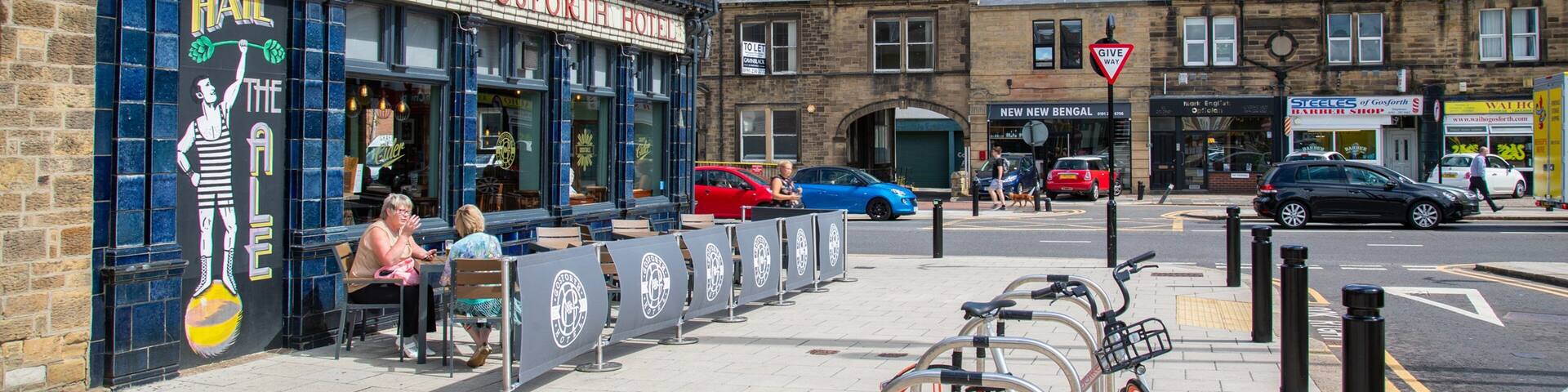A street with a pub named 'The Hail Ale' and 'The Gosforth Hotel'. There are bicycles parked on the sidewalk, a 'Give Way' sign, and cars on the road.