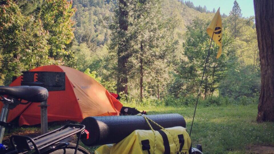 A red tent, a bicycle, and a yellow bag with a sign in a forested area.