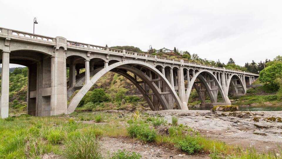 A concrete bridge with multiple arches spanning a river, surrounded by greenery and hills.