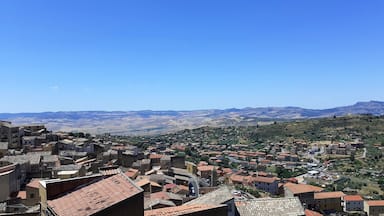 A town with terracotta-roofed buildings nestled in a valley, surrounded by rolling hills and mountains under a clear blue sky.