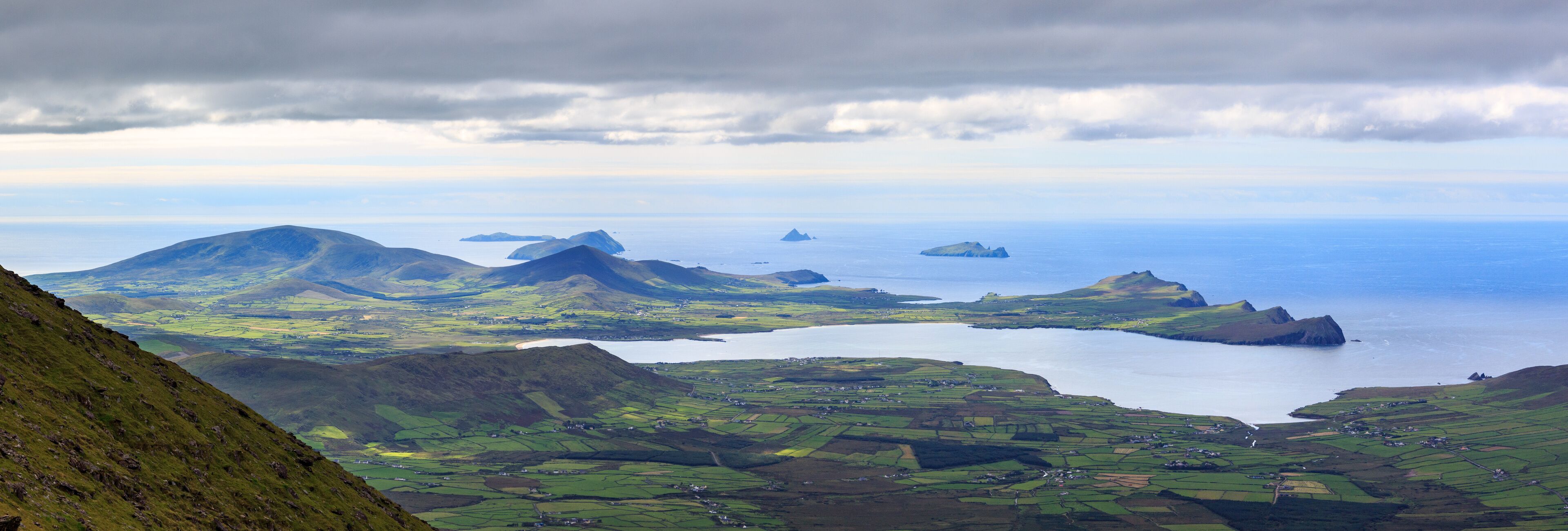 A coastal landscape with green fields, a lake, and distant islands under a cloudy sky.