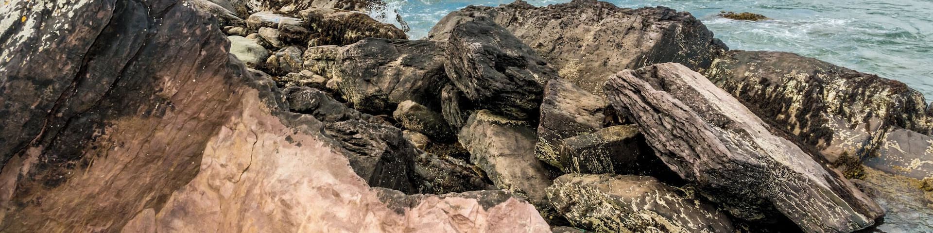 A rocky coastline with waves crashing against the rocks, a distant view of green hills, and a clear blue sky.
