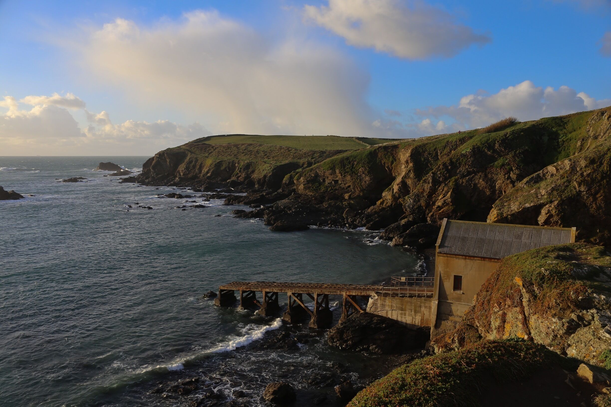 A coastal landscape with a bridge over the water, cliffs, and a small building on the right.