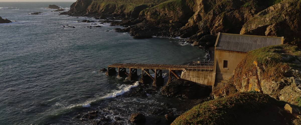 A coastal landscape with a bridge over the water, cliffs, and a small building on the right.