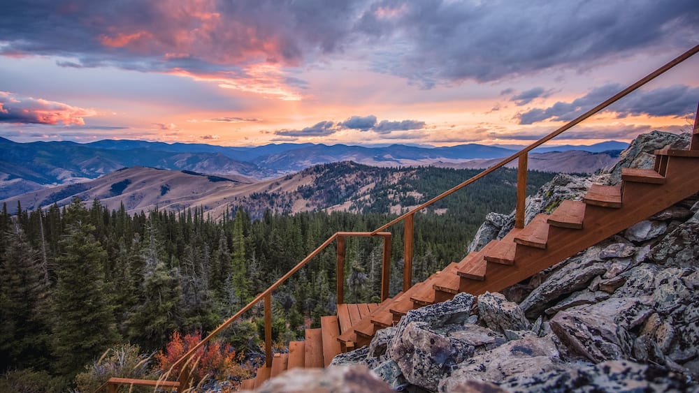 Un escalier en bois menant au sommet d’une pente montagneuse rocheuse, d’où l’on peut admirer des vallées verdoyantes et des montagnes lointaines, le tout baigné par les couleurs d’un coucher de soleil spectaculaire.