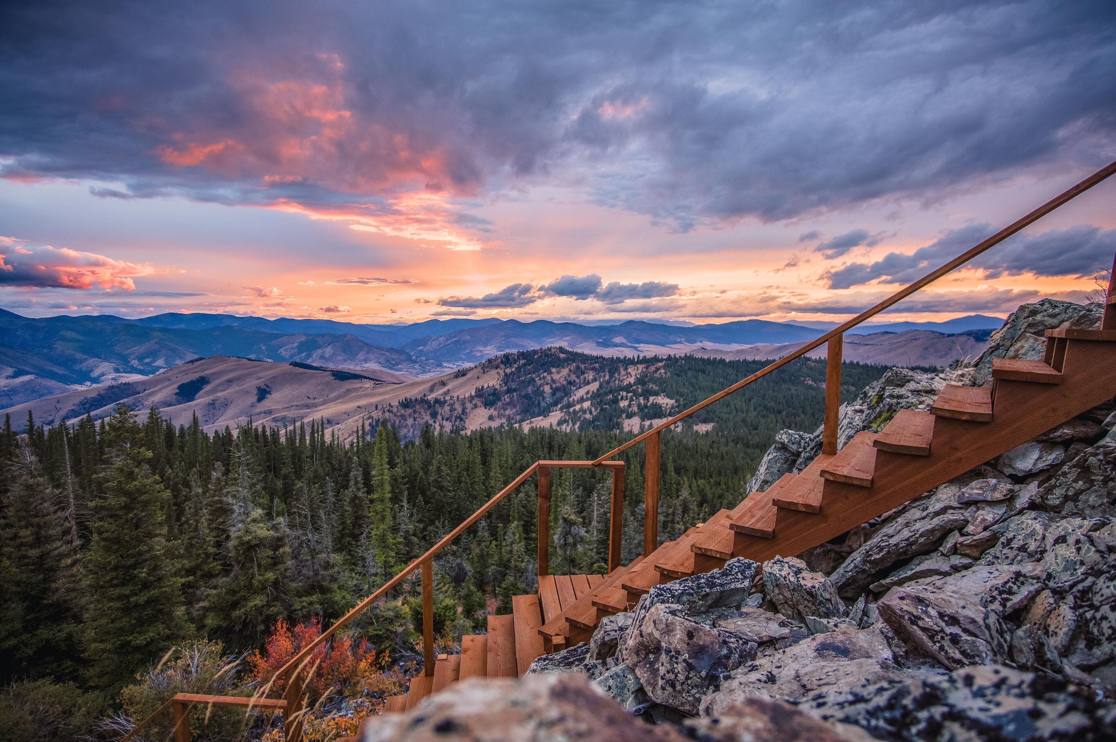 A wooden staircase leading up a rocky mountain slope with a view of forested valleys and distant mountains under a colorful sunset sky.