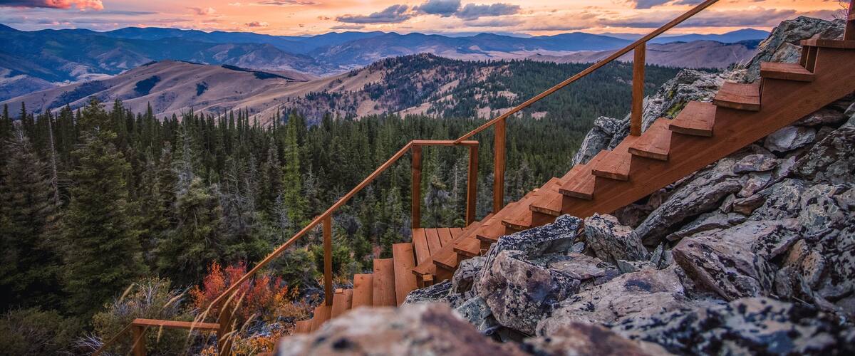 A wooden staircase leading up a rocky mountain slope with a view of forested valleys and distant mountains under a colorful sunset sky.