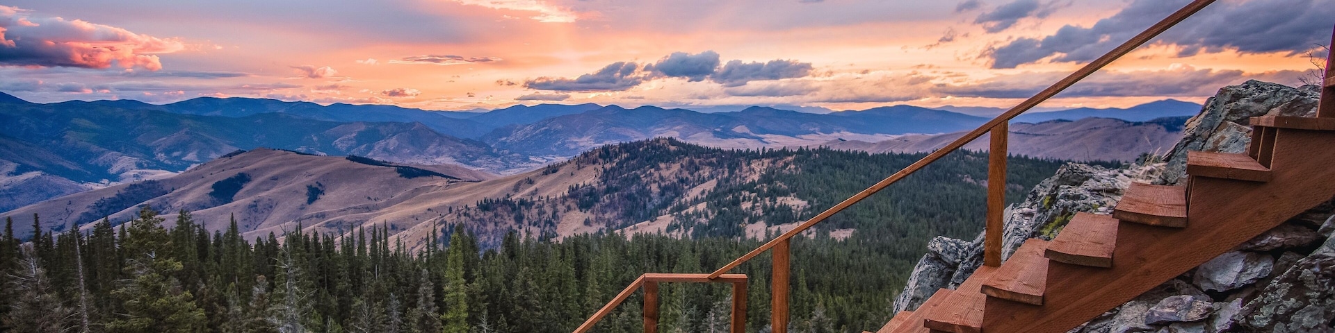 A wooden staircase leading up a rocky mountain slope with a view of forested valleys and distant mountains under a colorful sunset sky.