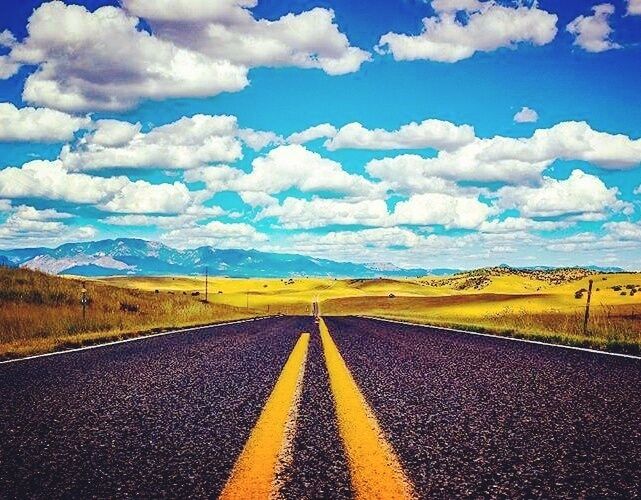 A road with yellow center lines, surrounded by grassy fields and mountains under a blue sky with clouds.