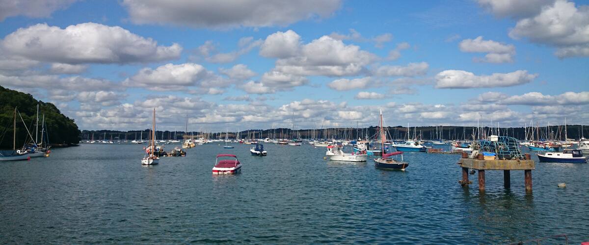 A marina with various boats docked, a clear sky with scattered clouds, and a forested hill in the background.