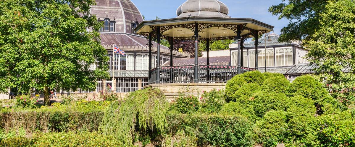 A gazebo surrounded by lush greenery, a large building with a dome, and a flagpole with a flag.