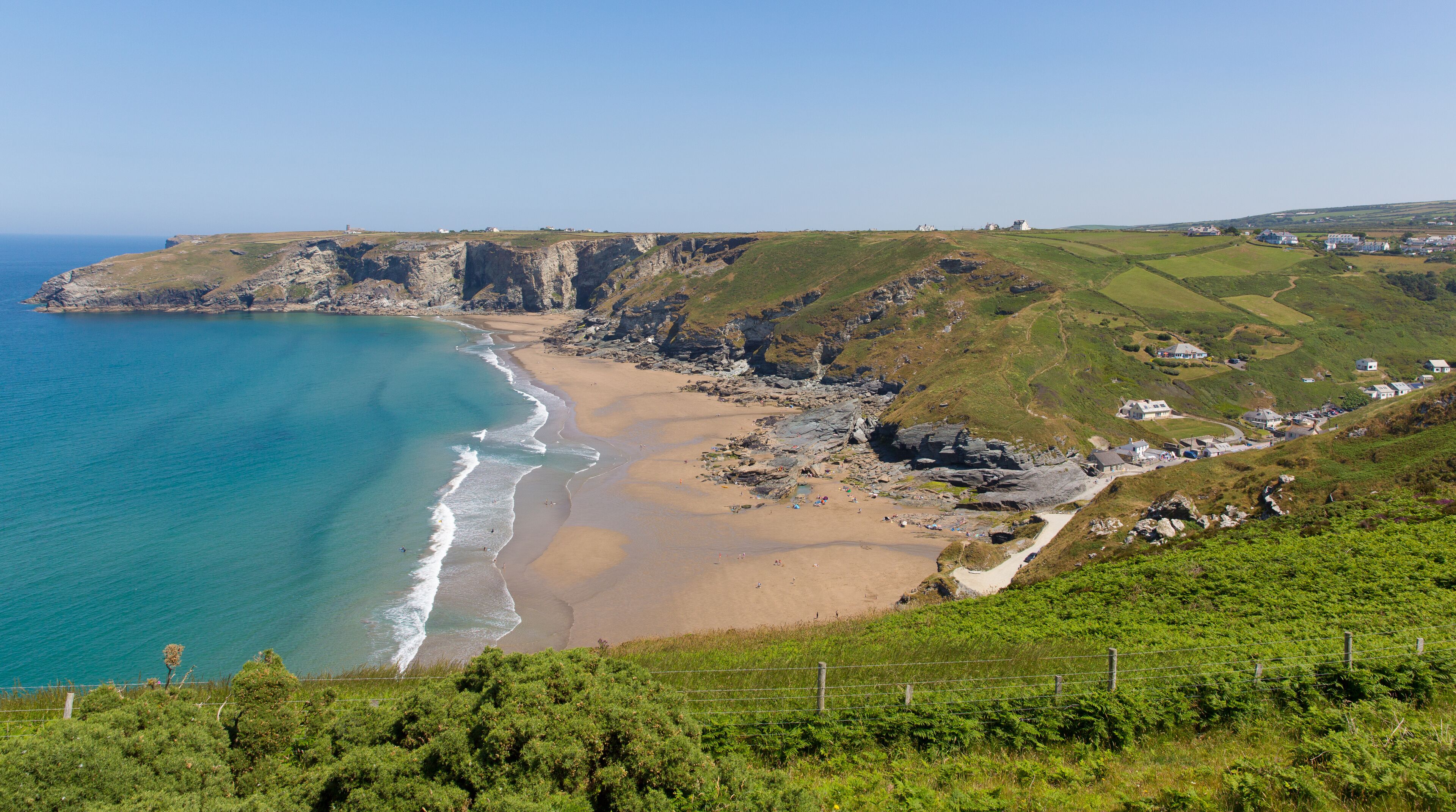 A coastal landscape with a sandy beach, rocky cliffs, and a grassy hill with houses.
