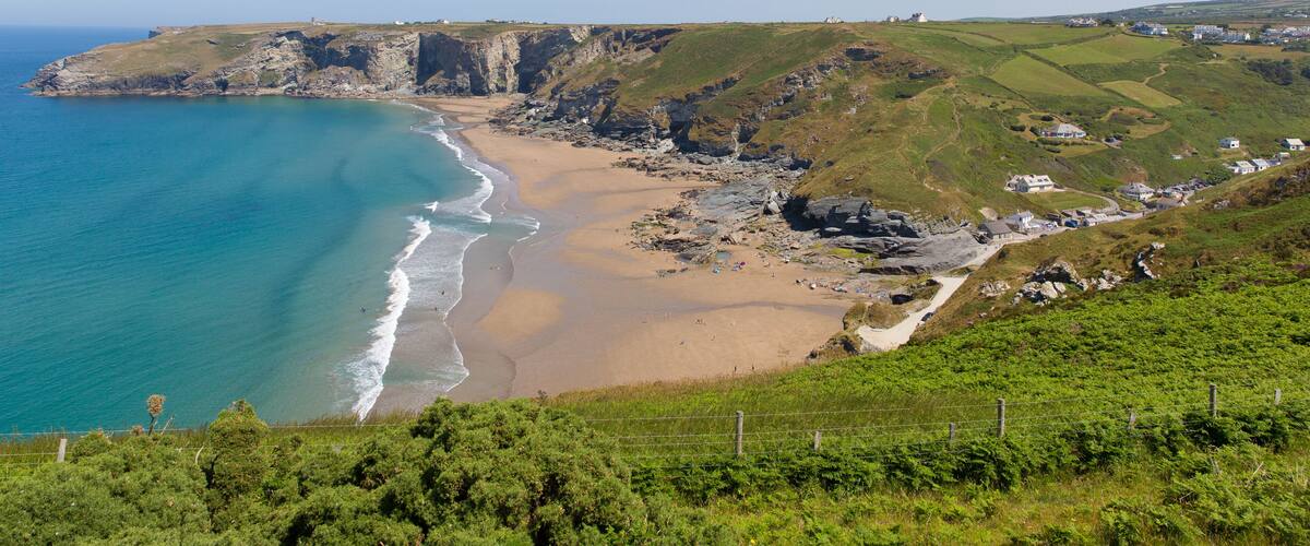 A coastal landscape with a sandy beach, rocky cliffs, and a grassy hill with houses.