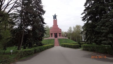Um monumento com uma estátua no topo, cercado por árvores e um caminho pavimentado.