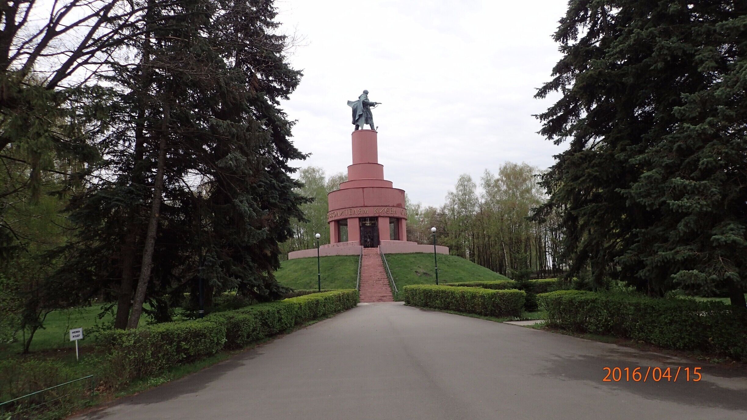 A monument with a statue on top, surrounded by trees and a paved path.