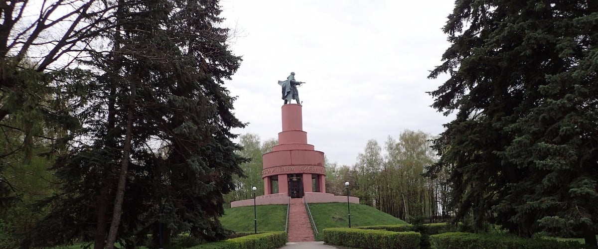 A monument with a statue on top, surrounded by trees and a paved path.