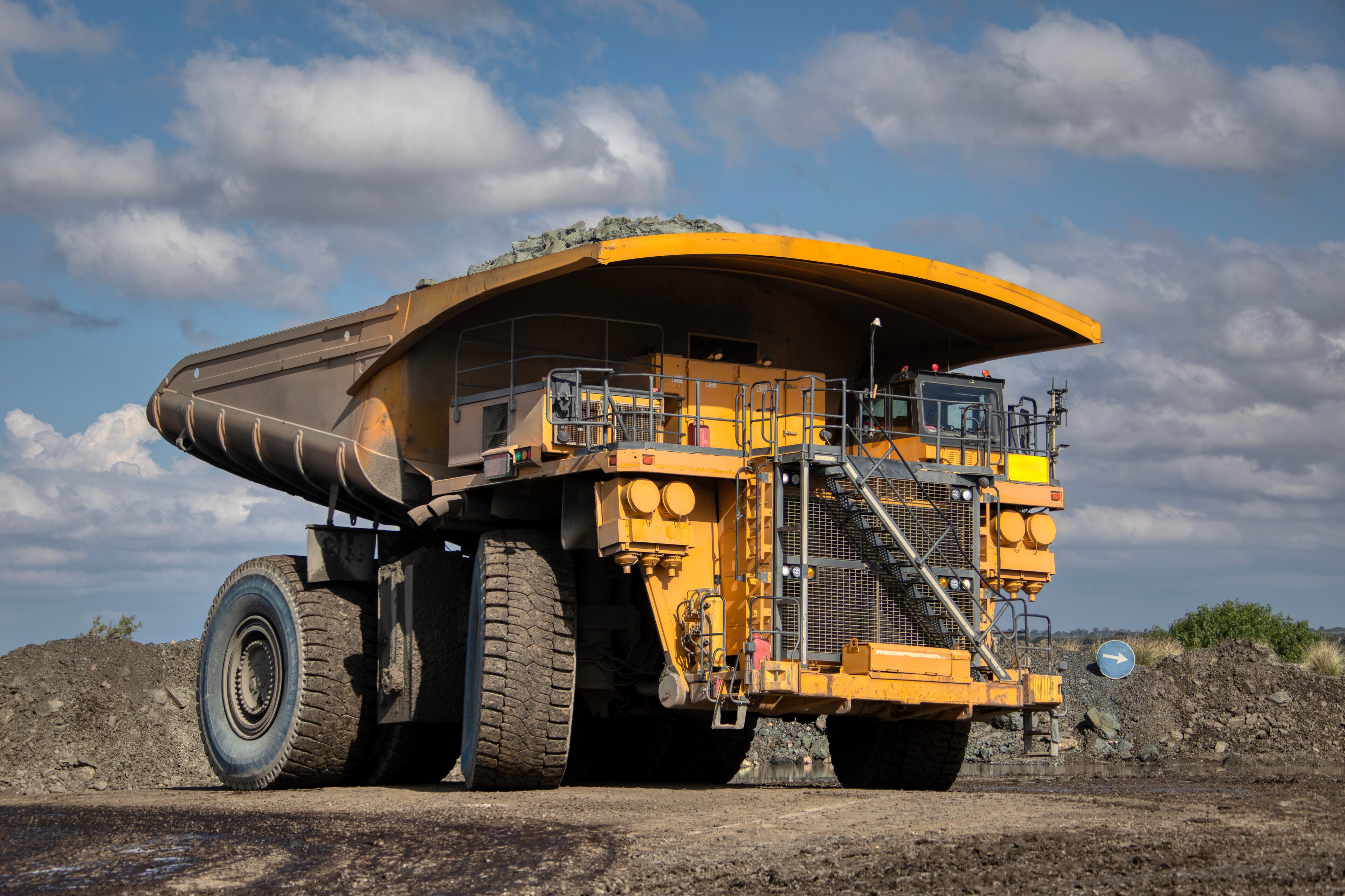 A large yellow dump truck with a curved bed, parked on a dirt road with a rocky hill in the background and a partly cloudy sky.