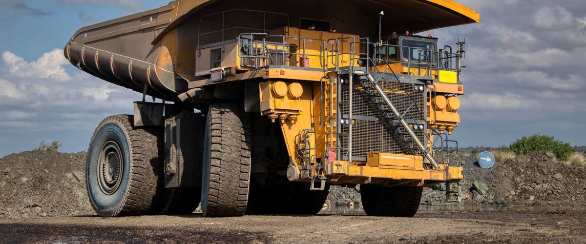 A large yellow dump truck with a curved bed, parked on a dirt road with a rocky hill in the background and a partly cloudy sky.