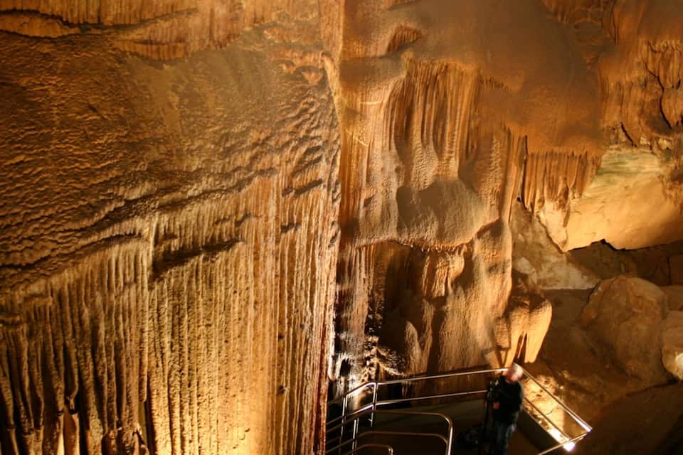 A cave with stalactites and stalagmites, a metal railing, and a person standing near the railing.
