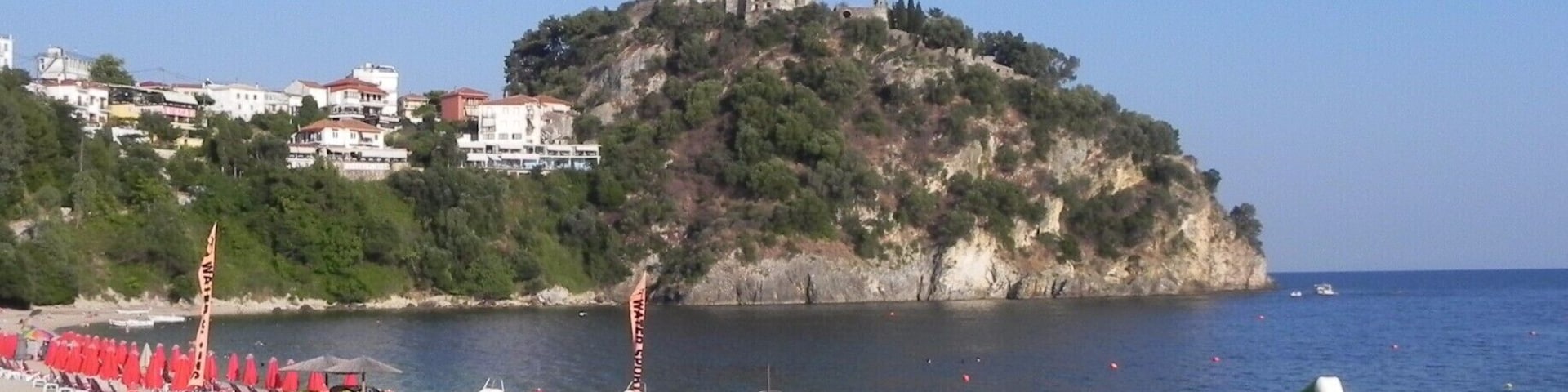 Una playa con sombrillas rojas y tumbonas, una colina rocosa con un castillo al fondo, y un mar tranquilo.
