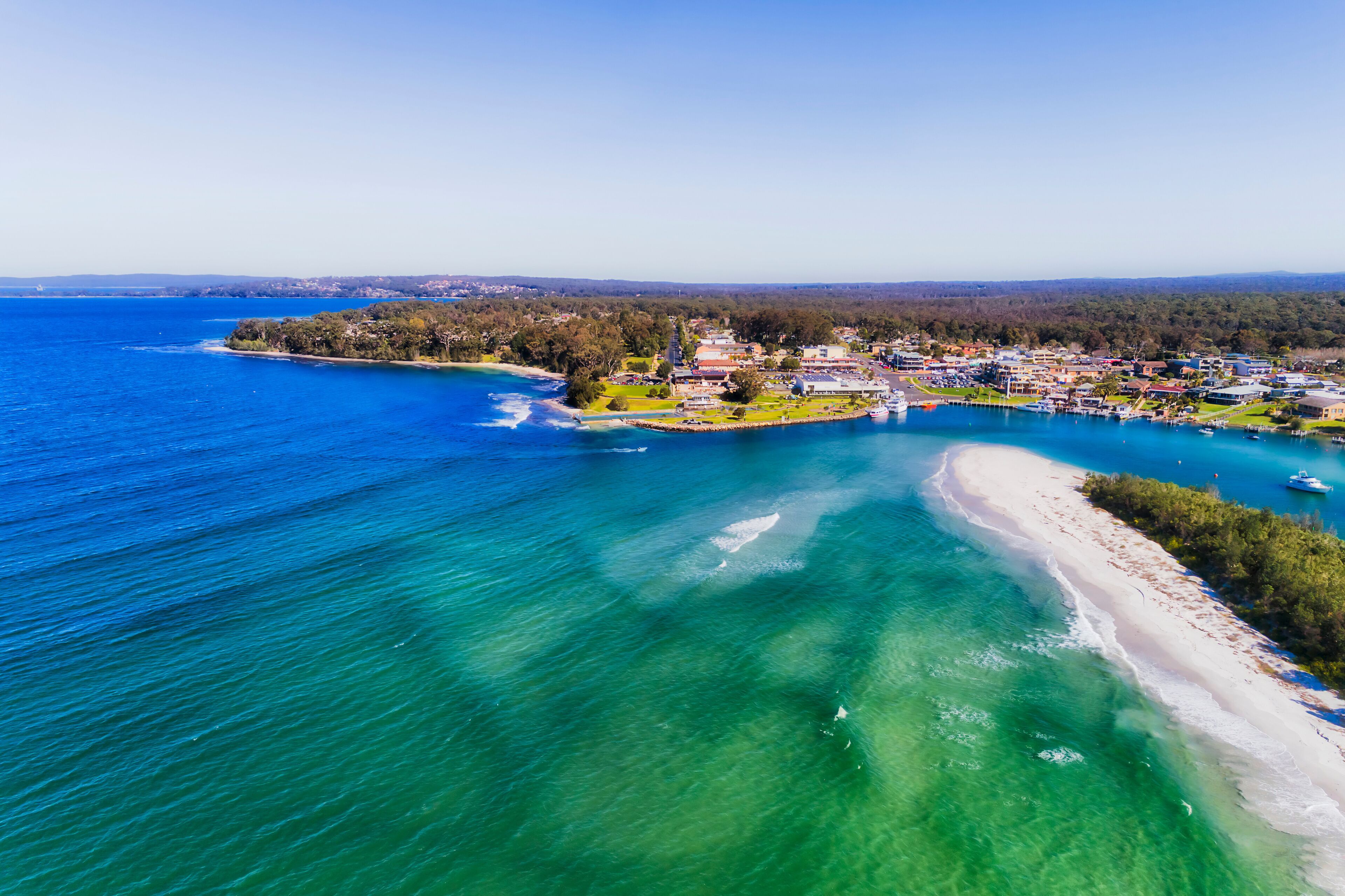 An aerial view of a coastal town with a beach, clear blue waters, and surrounding greenery.