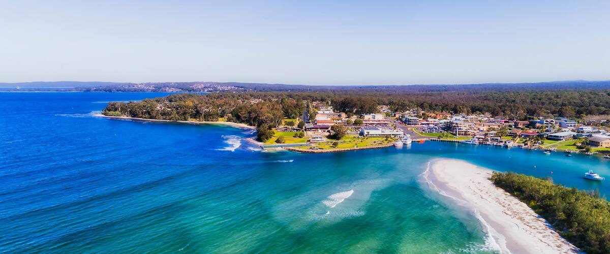 An aerial view of a coastal town with a beach, clear blue waters, and surrounding greenery.