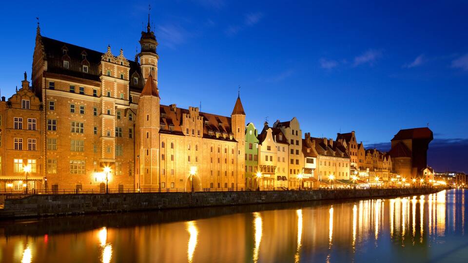 A historic waterfront cityscape at dusk with illuminated buildings reflecting on the water.