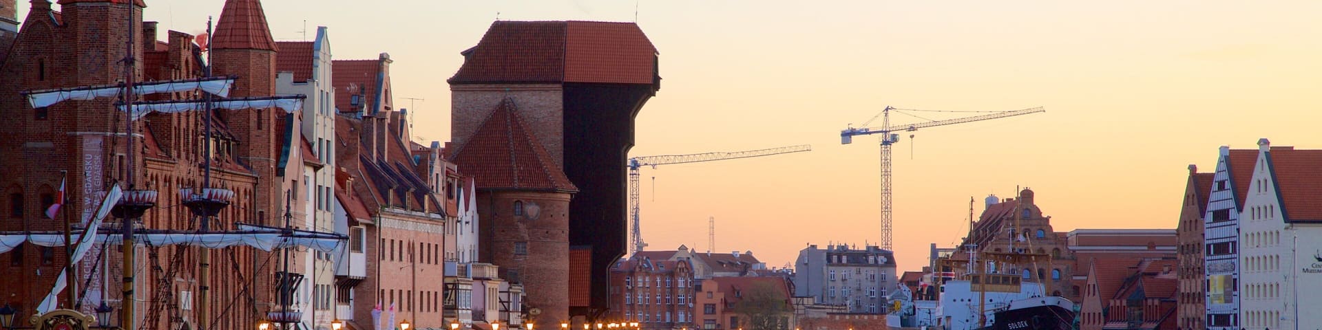 A historic waterfront with traditional European architecture, a large ship docked, and construction cranes in the background.