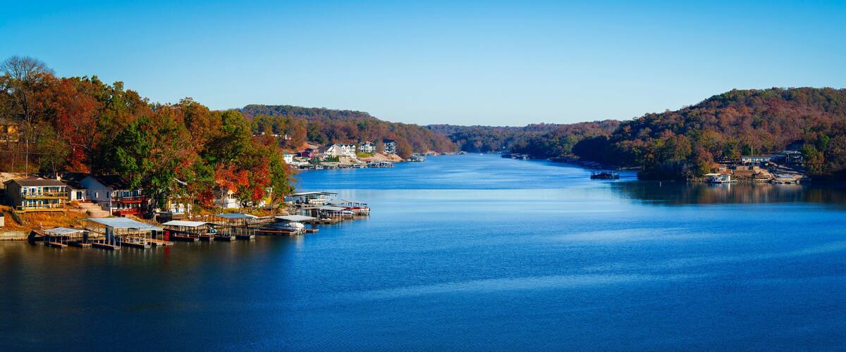 A calm lake with docked boats, surrounded by autumn-colored trees and houses on the shore.