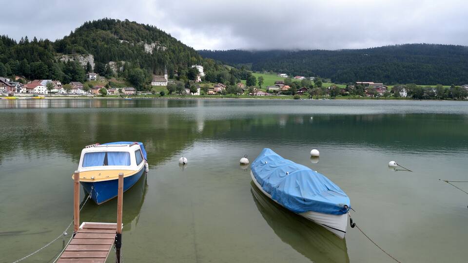 A lake with two boats docked, a wooden pier, and houses in the background.