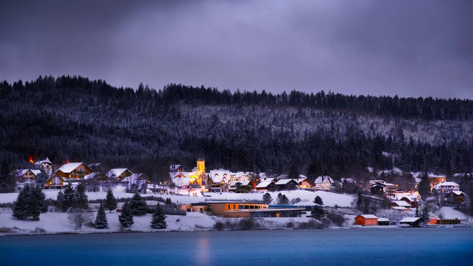 A snow-covered village by a lake at dusk with illuminated houses and a church tower.
