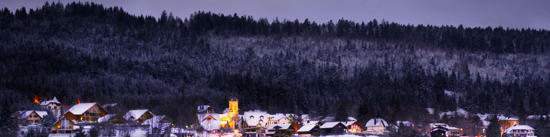 Un pueblo cubierto de nieve junto a un lago al anochecer, con casas iluminadas y una torre de iglesia.