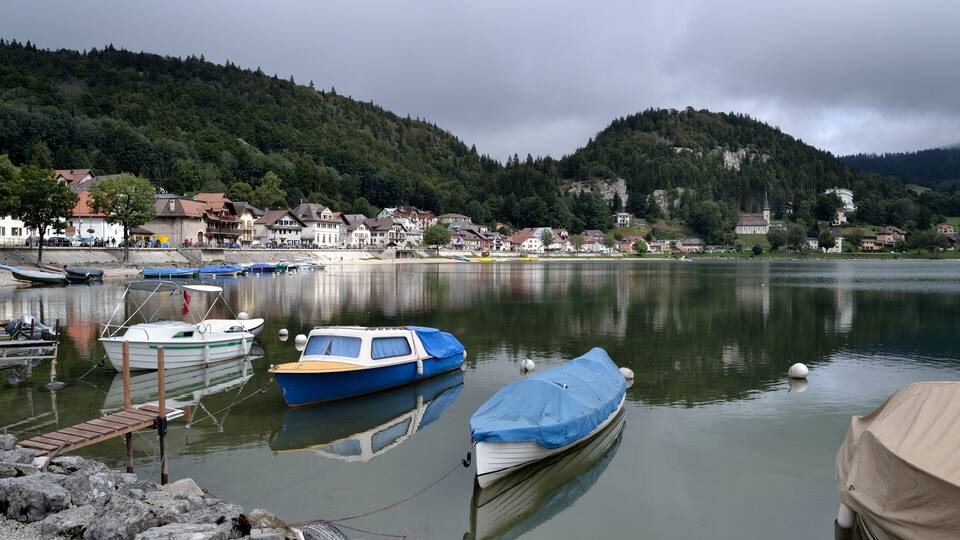 A calm lake with boats docked at the shore, surrounded by houses and hills.