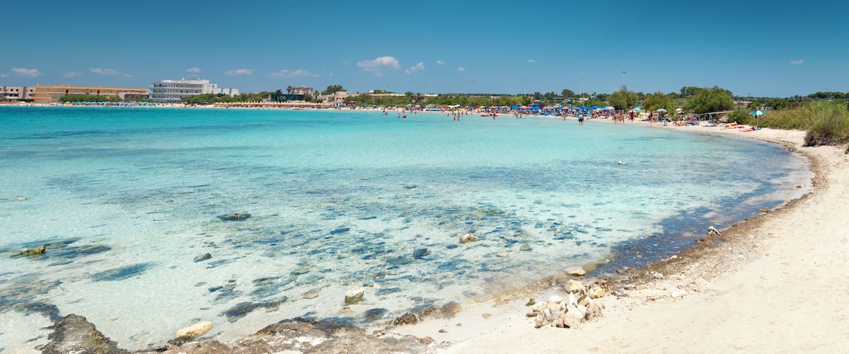 A beach with clear turquoise water, sandy shore, and a few buildings in the background.