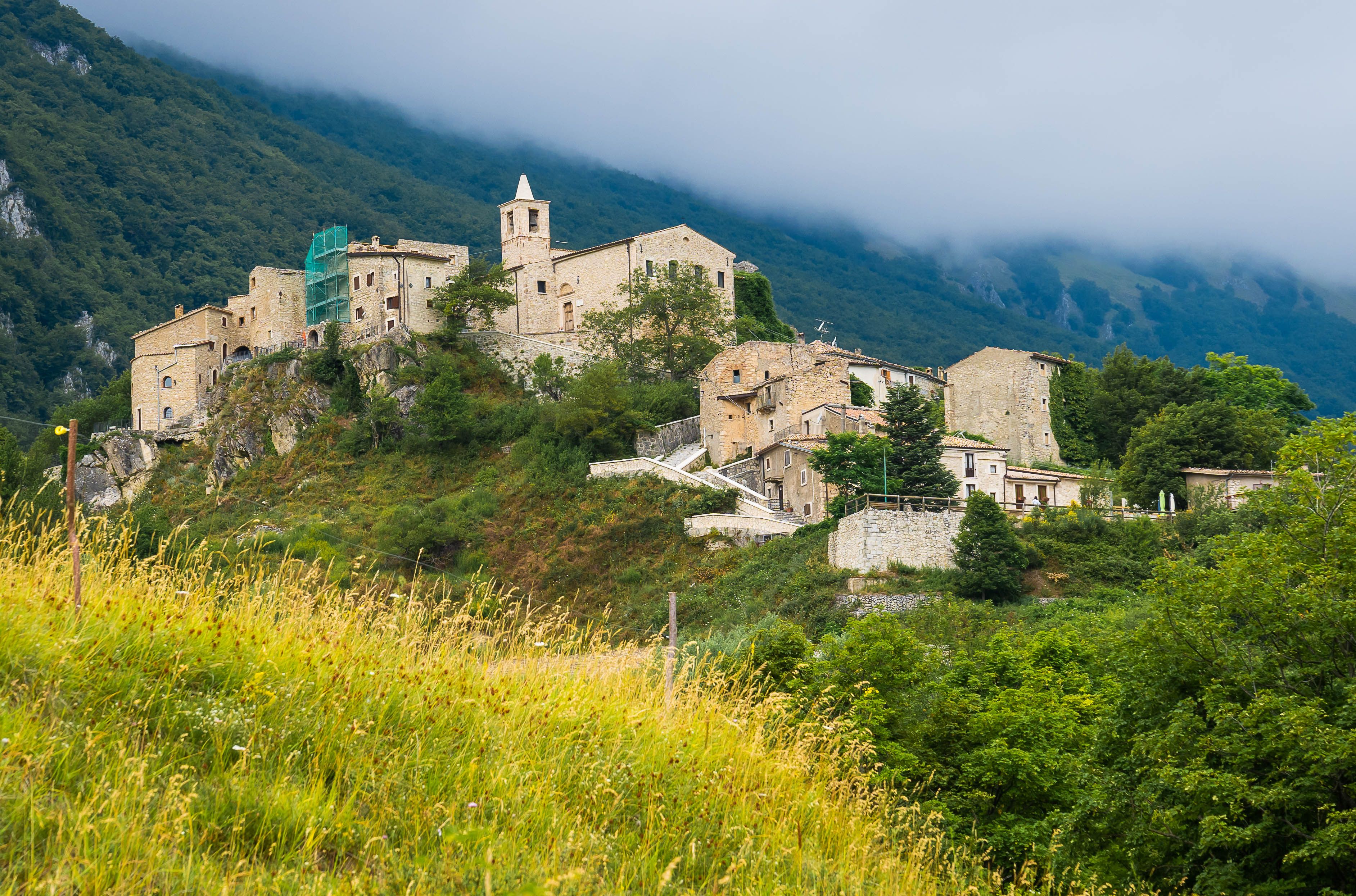 A historic stone village with a prominent church and tower, surrounded by lush greenery and mountains.