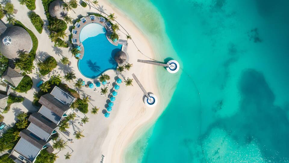 An aerial view of a resort with a swimming pool, beach, and palm trees.