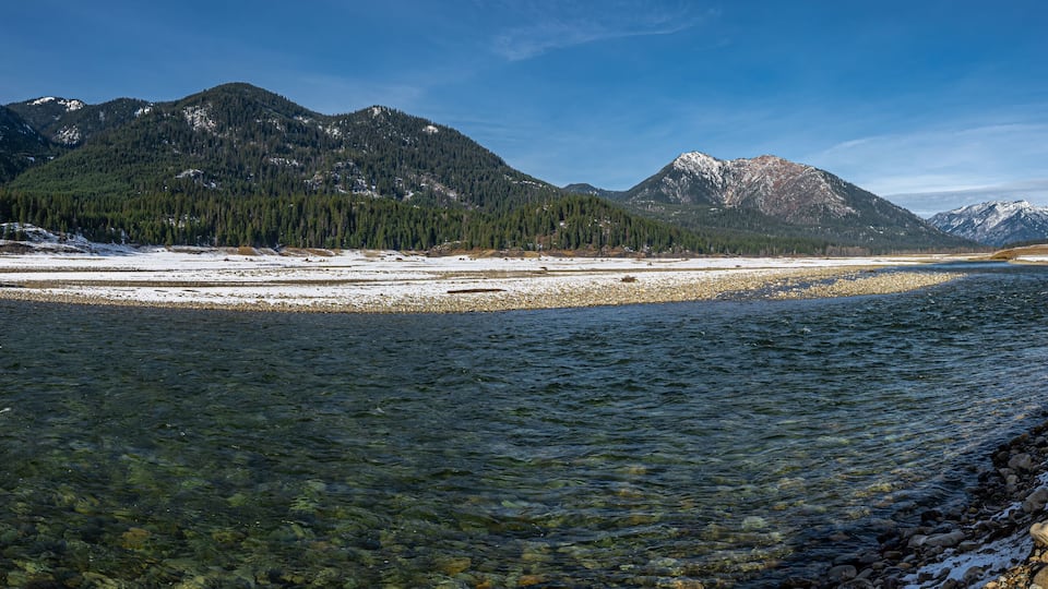 A clear river with snow-covered banks and mountains in the background.