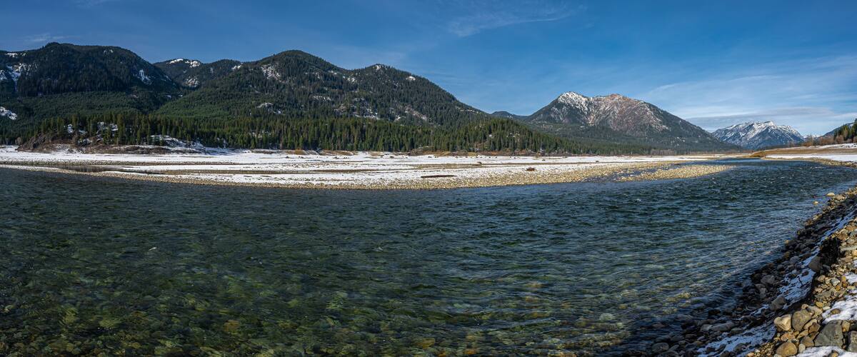 A clear river with snow-covered banks and mountains in the background.