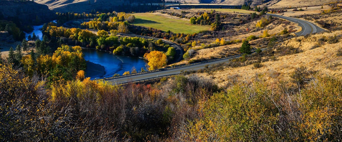 Une route sinueuse traverse une vallée où coule une rivière, le tout étant bordé de collines et d’arbres aux couleurs automnales.
