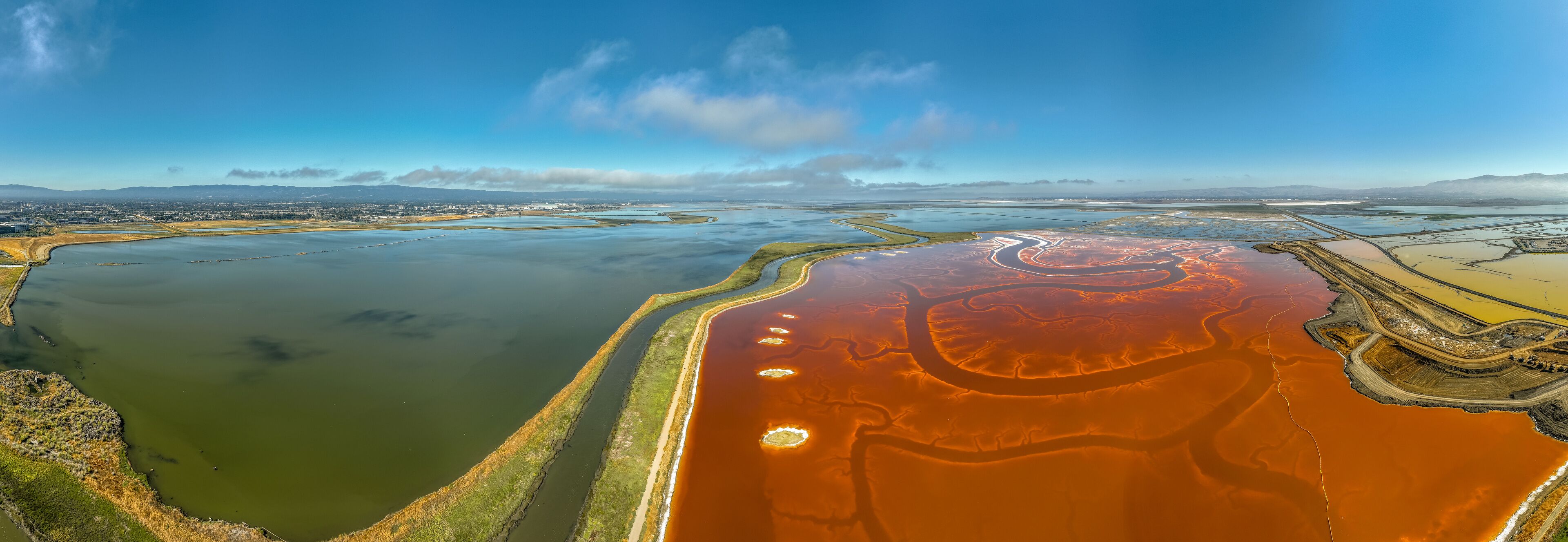 An aerial view of a large body of water with distinct red and green areas, surrounded by land and distant mountains.