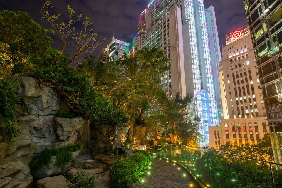 A nighttime cityscape with illuminated buildings, a stone pathway, and lush greenery.