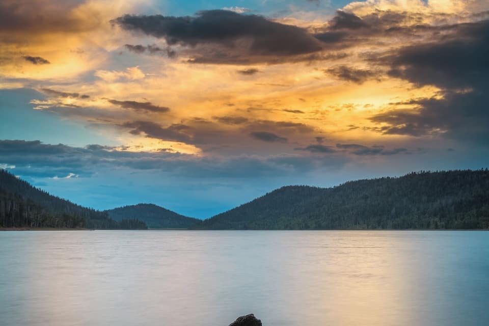 A calm lake with a small island in the center, surrounded by forested hills under a dramatic sky.