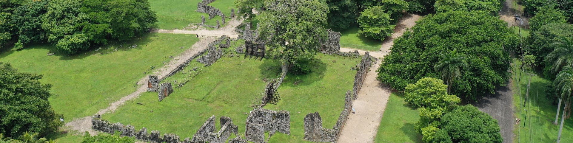 An aerial view of a historical site with ruins, dense greenery, and a coastal backdrop with a bridge.