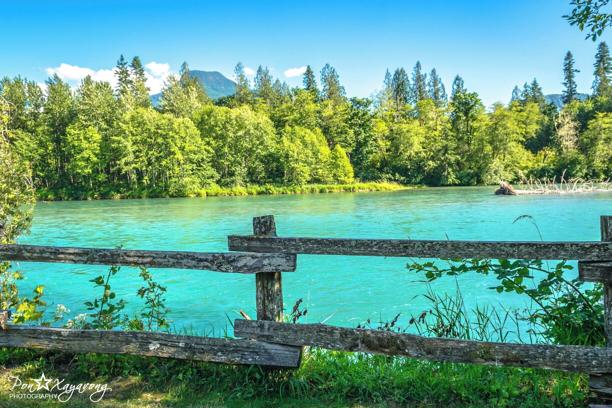 A turquoise river with a wooden fence in the foreground, surrounded by dense green forest and mountains in the background.