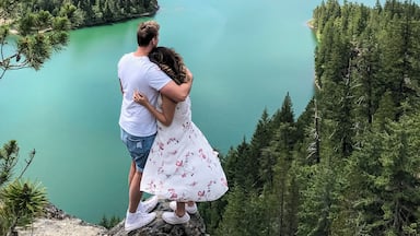 A couple embracing on a rocky outcrop overlooking a turquoise lake surrounded by dense forests and snow-capped mountains.